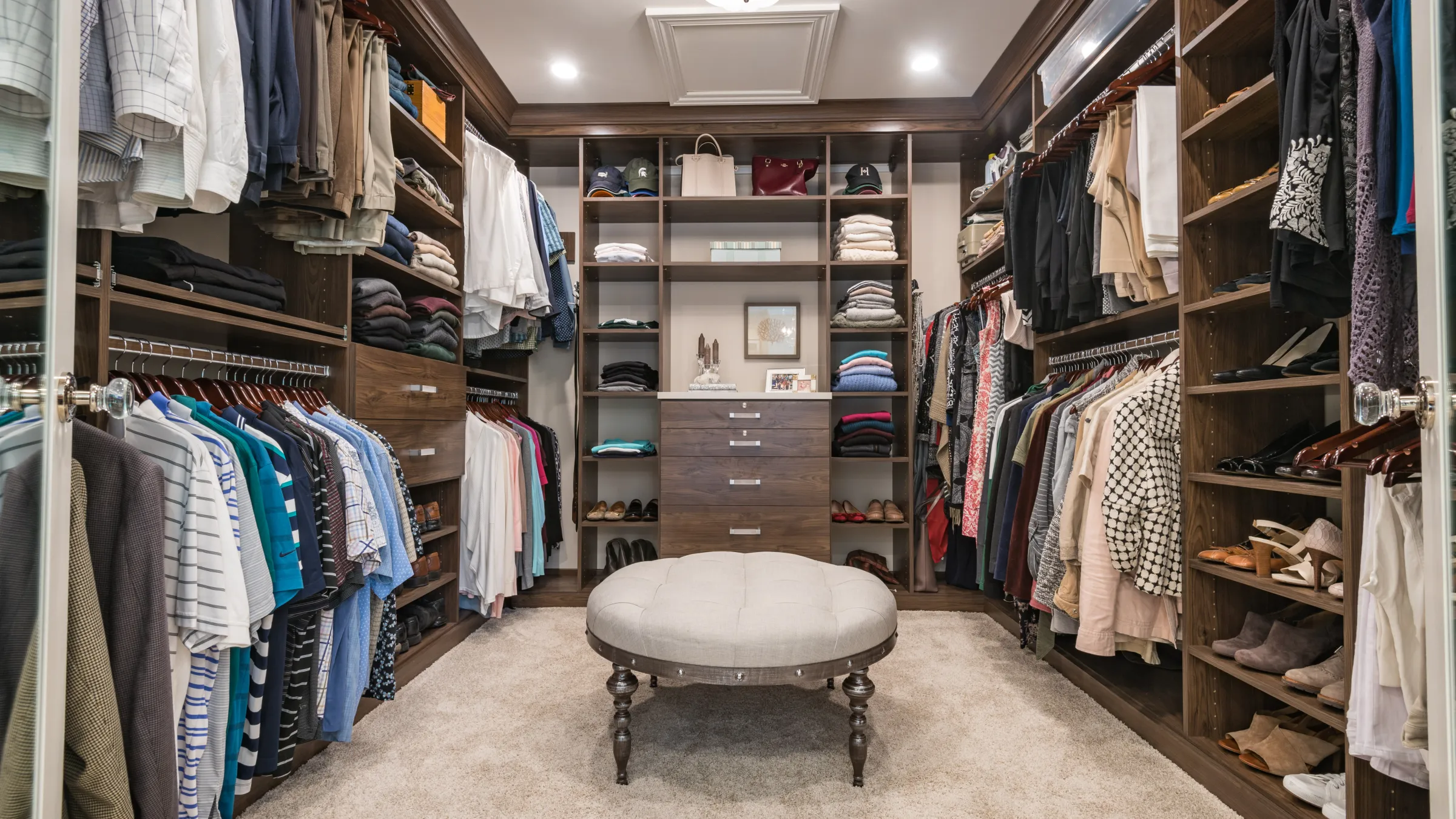 Custom walk-in closet with dark wood cabinetry, shelving, ottoman, and organized clothing in Palatine, IL.