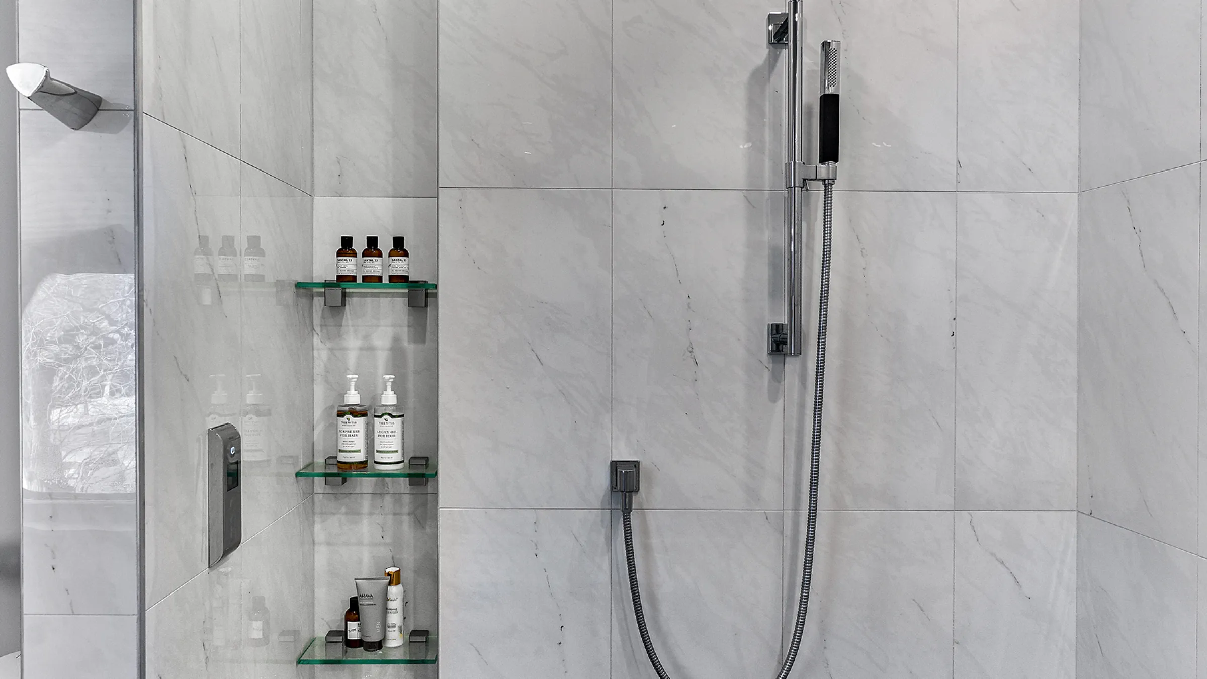 Modern bathroom shower with marble tile, glass shelves, and handheld shower head.