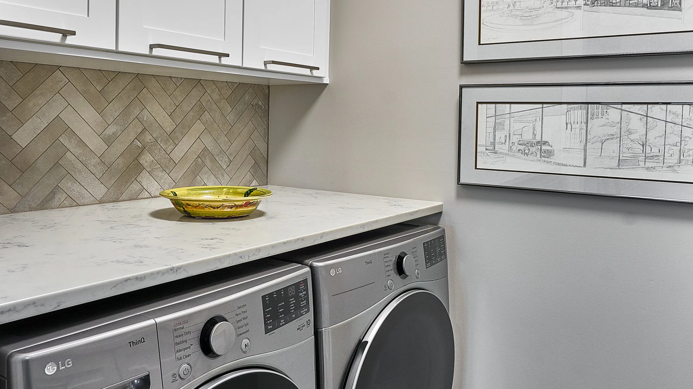 Modern laundry room featuring LG washer/dryer, white cabinets, herringbone backsplash, and light marble countertop.