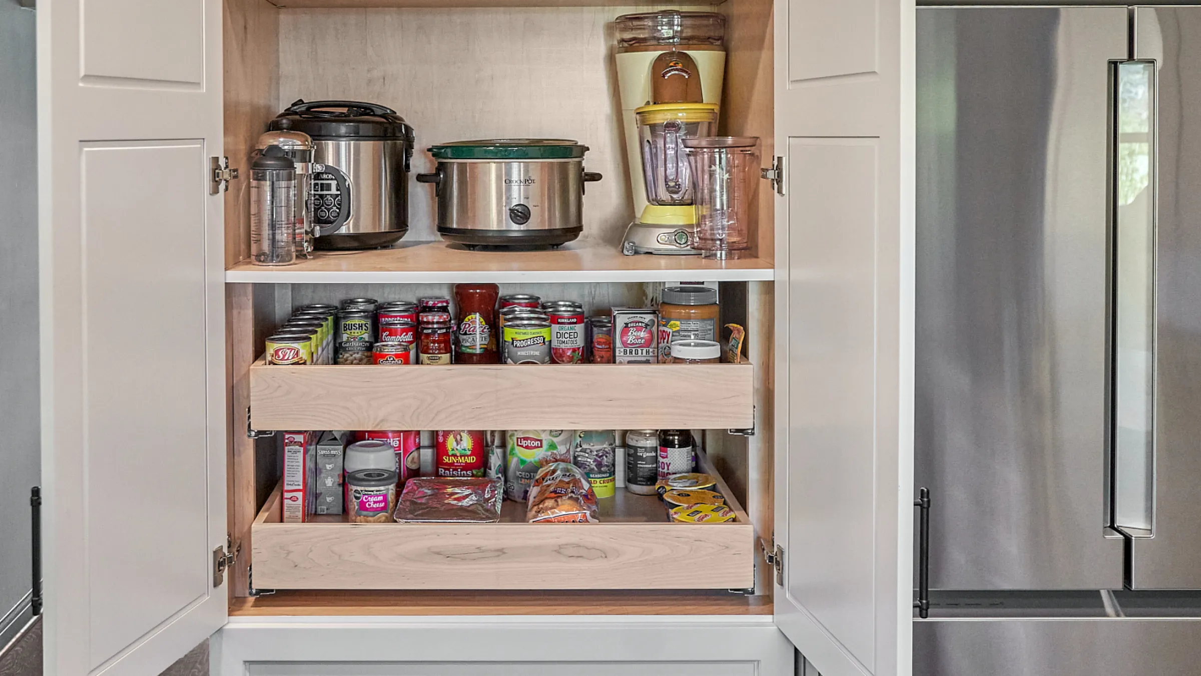 Open kitchen pantry with pull-out shelves stocked with canned goods and appliances. White cabinets are visible.