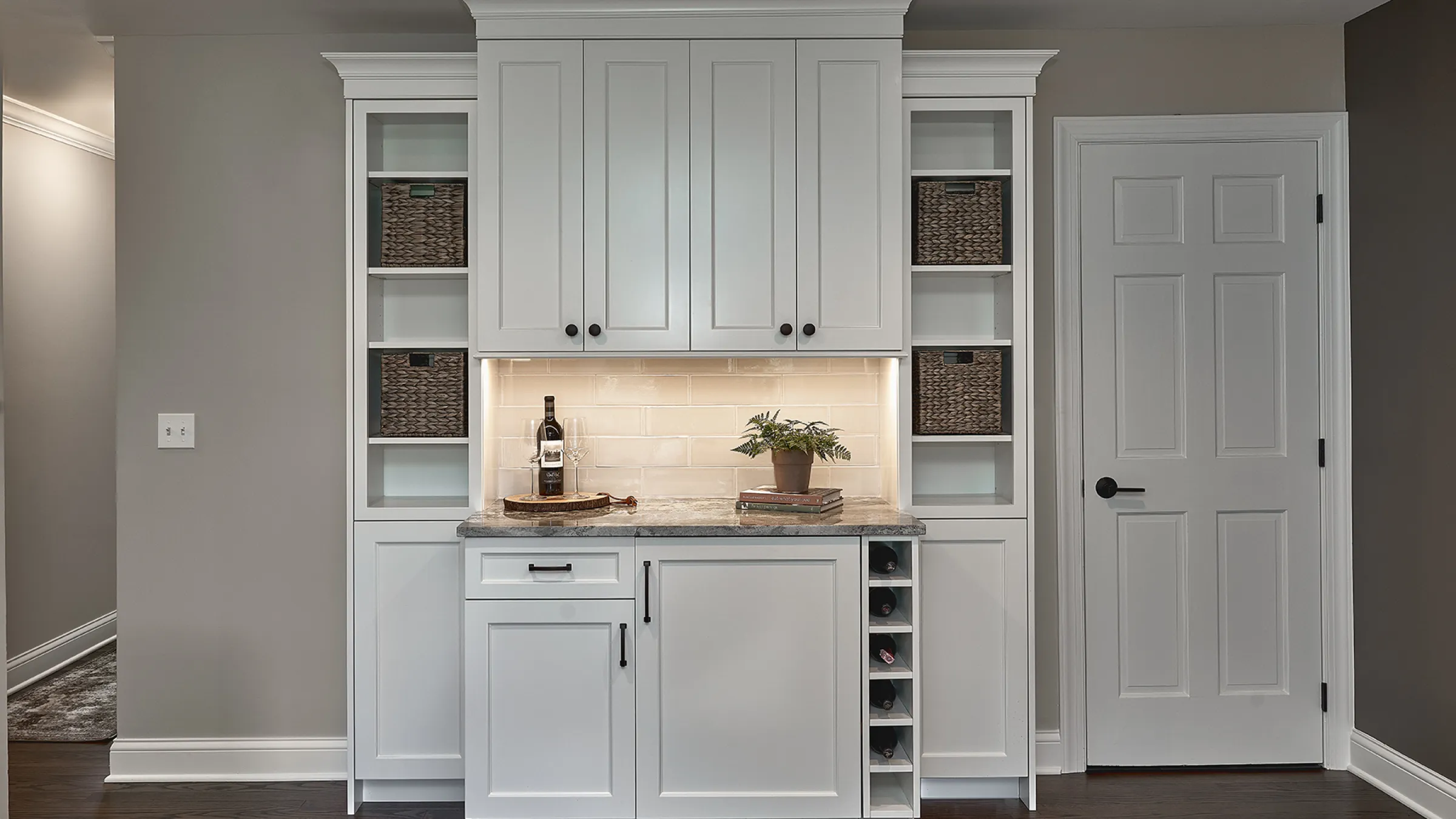 Custom white kitchen cabinetry with wine rack, granite countertops, woven baskets, and recessed lighting.