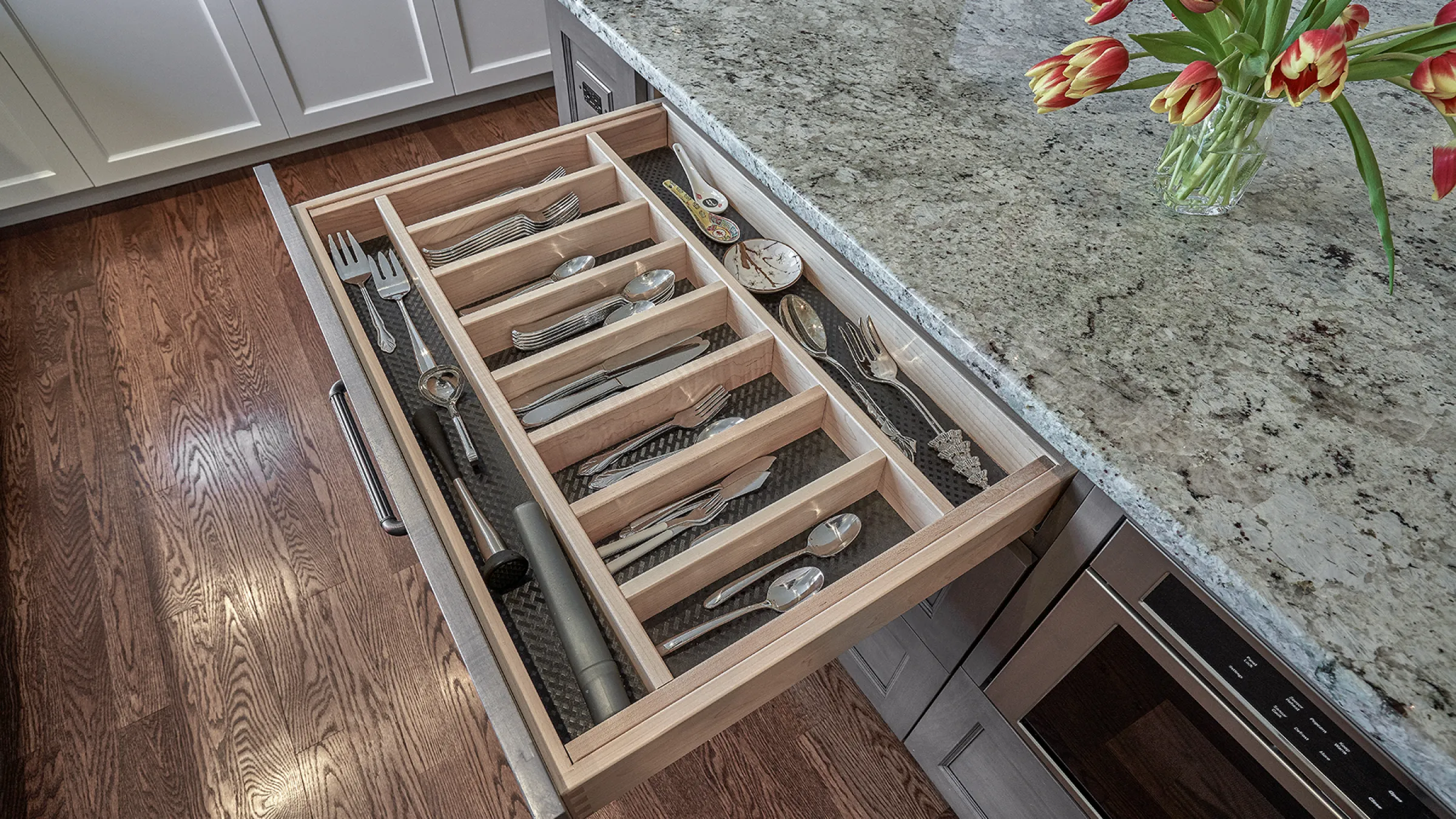 Open kitchen drawer with custom wood dividers organizing silverware and utensils. Granite countertop and hardwood floors.