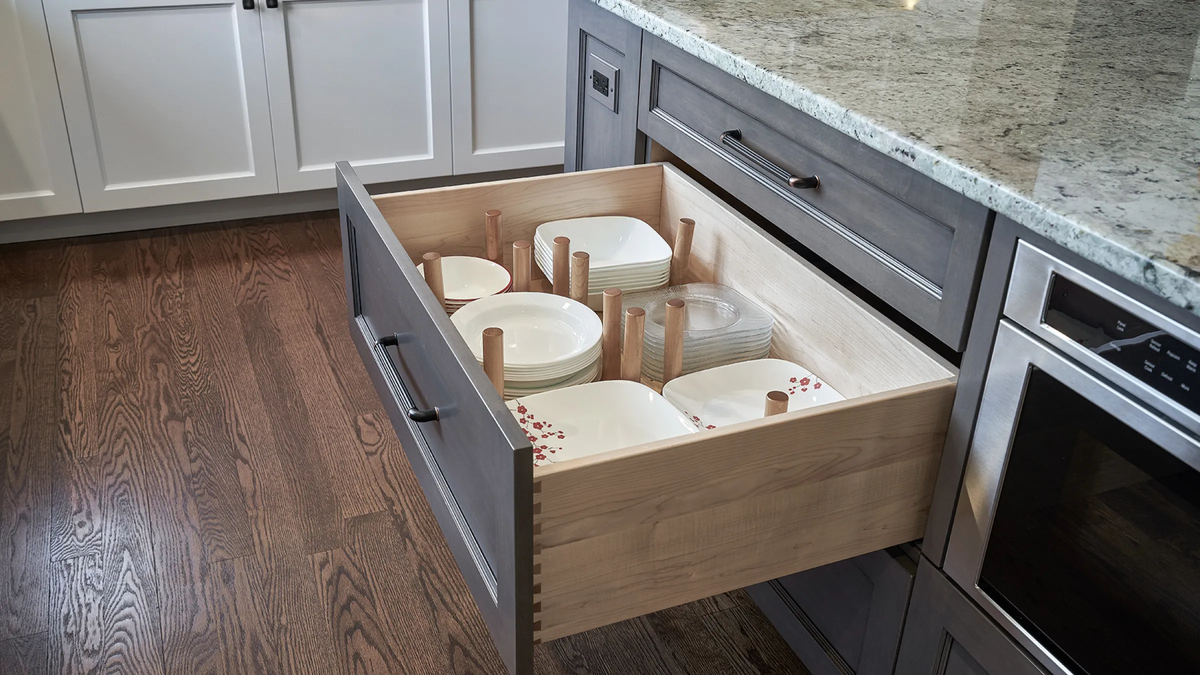 Open kitchen drawer with dish organization pegs. Gray cabinets, granite countertops, and hardwood floors.