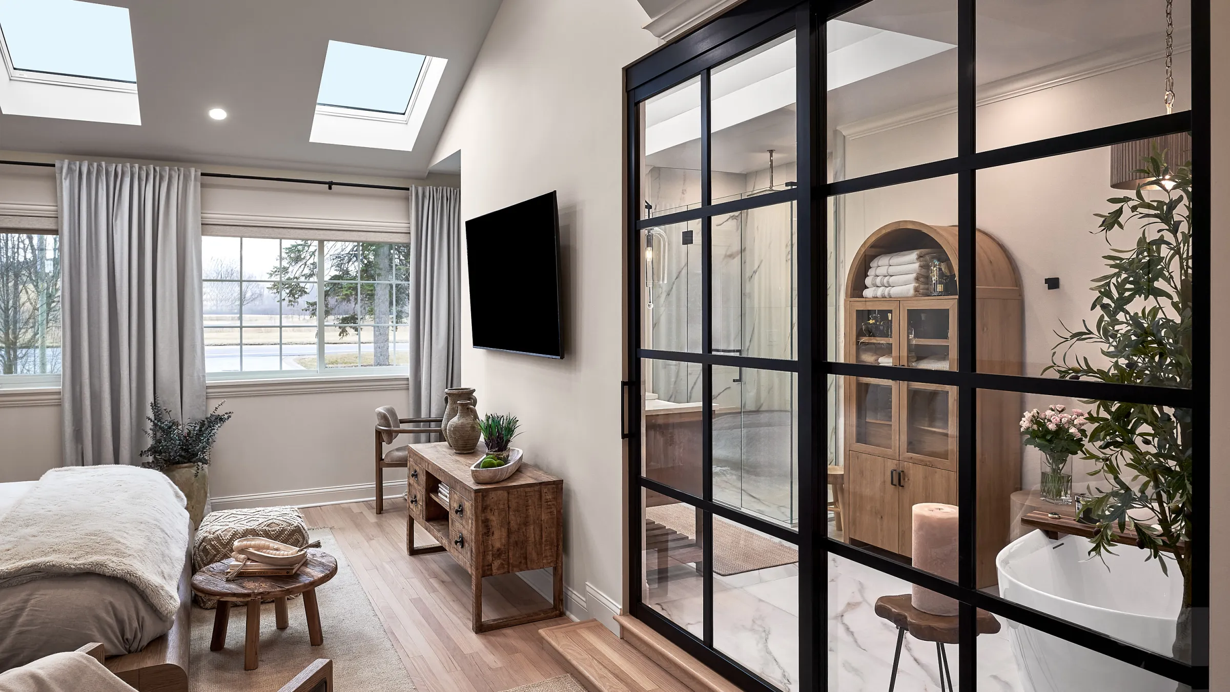 Modern bedroom with glass-enclosed bath featuring wood cabinet, marble floor, and soaking tub. Skylights provide natural ligh