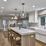 Modern white kitchen with large island, brass globe pendants, white shaker cabinets, farmhouse sink, and wood floors.
