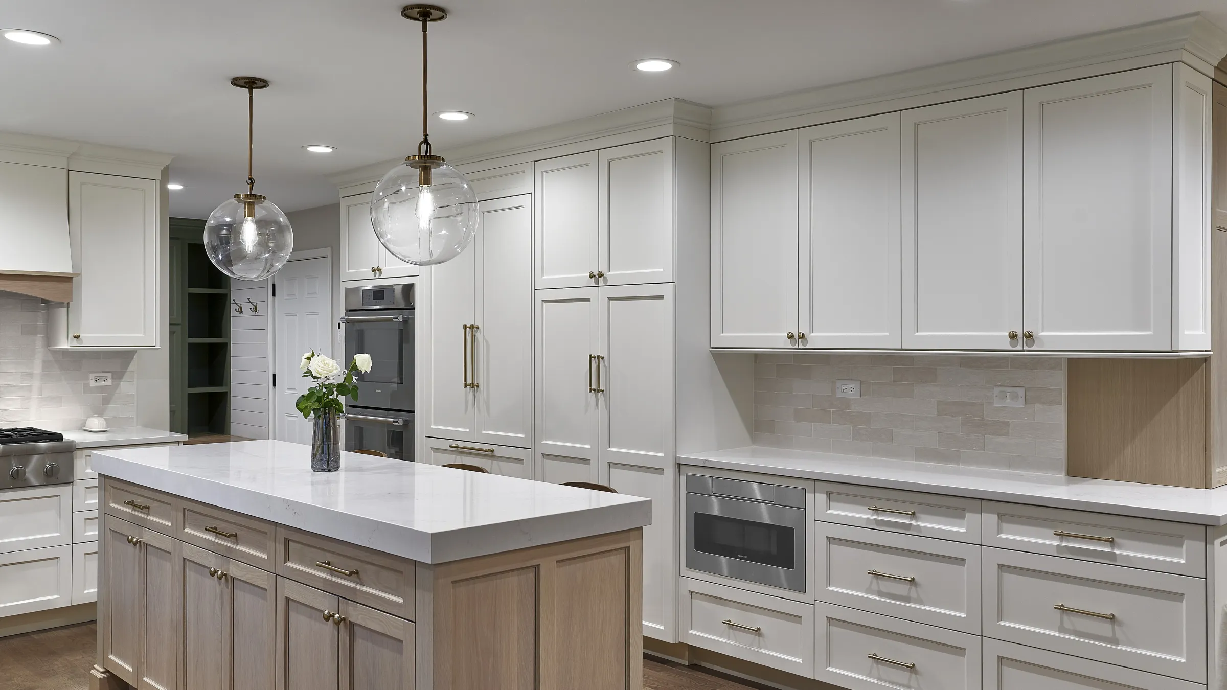 Modern white kitchen with large island, brass globe pendants, built-in appliances, light wood cabinets & white quartz counter