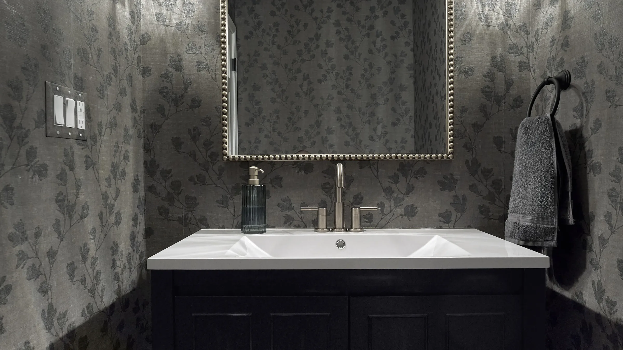 Modern powder room with dark floral wallpaper, white vanity top, dark blue cabinet, brushed nickel faucet, and towel ring.