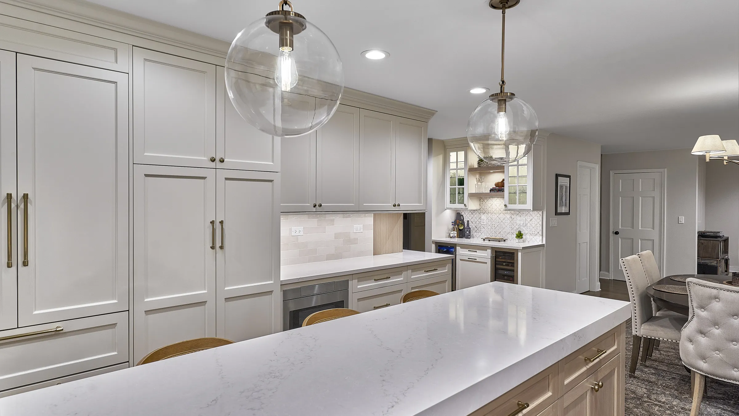 Modern white kitchen with large island, shaker cabinets, brass hardware, pendant lights, and light wood dining chairs.