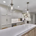 Modern white kitchen with large island, shaker cabinets, brass hardware, pendant lights, and light wood dining chairs.