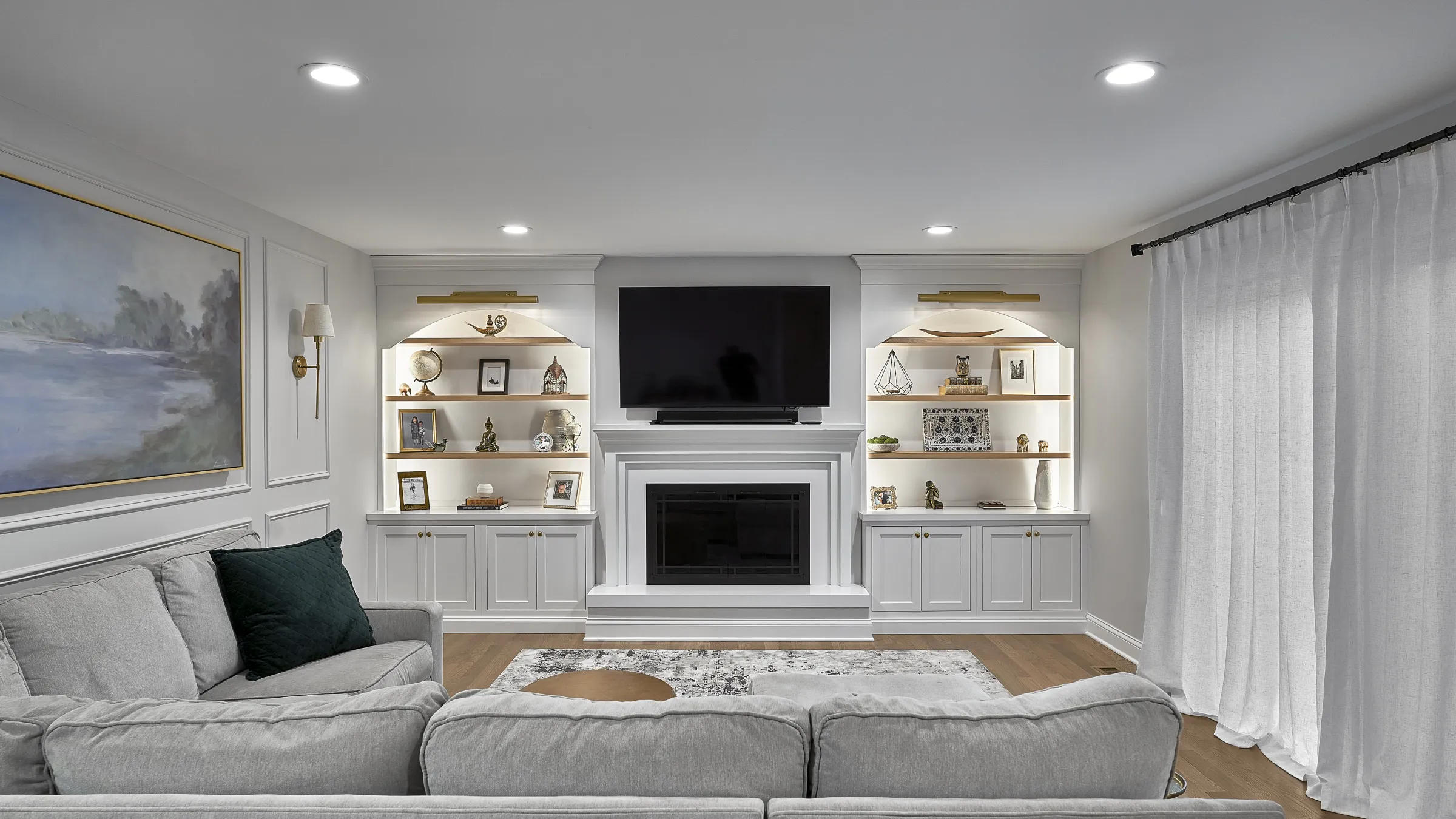 Modern living room with white built-in entertainment center, fireplace, recessed lighting, light gray sofa, and sheer white c