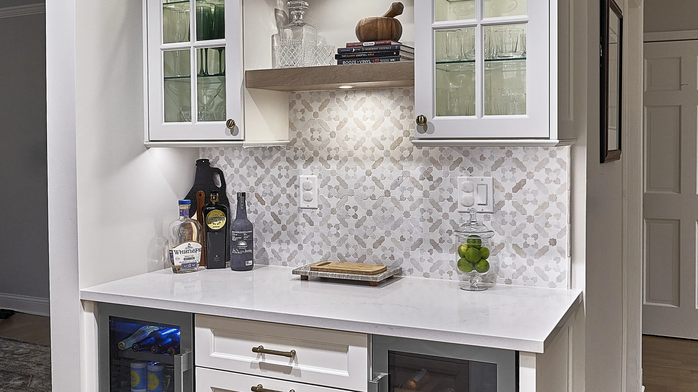Wet bar with white shaker cabinets, quartz countertop, patterned tile backsplash, beverage fridges, and liquor bottles.