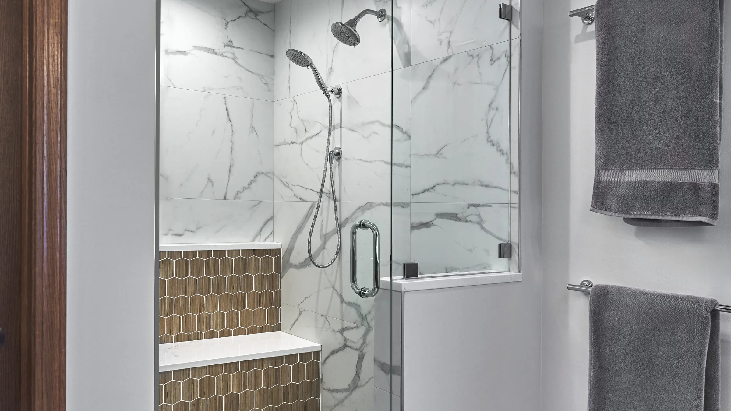 Modern primary bathroom shower with white marble-look tiles, a built-in bench with hexagonal wood-look tiles, and chrome fixt