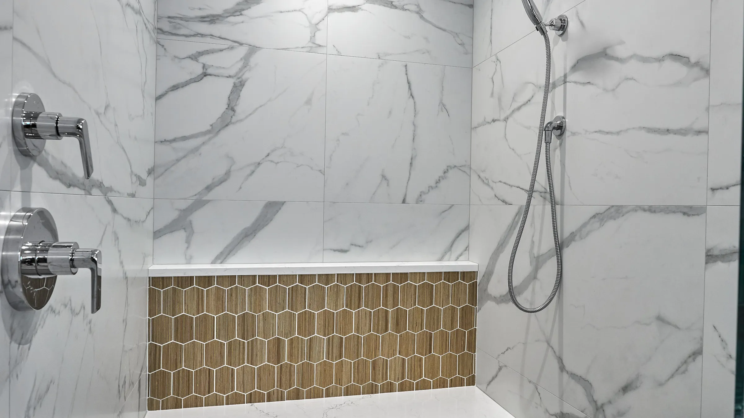 Modern primary bathroom shower with white marble-look tiles, chrome fixtures, and a wood-look hexagonal tile accent niche.