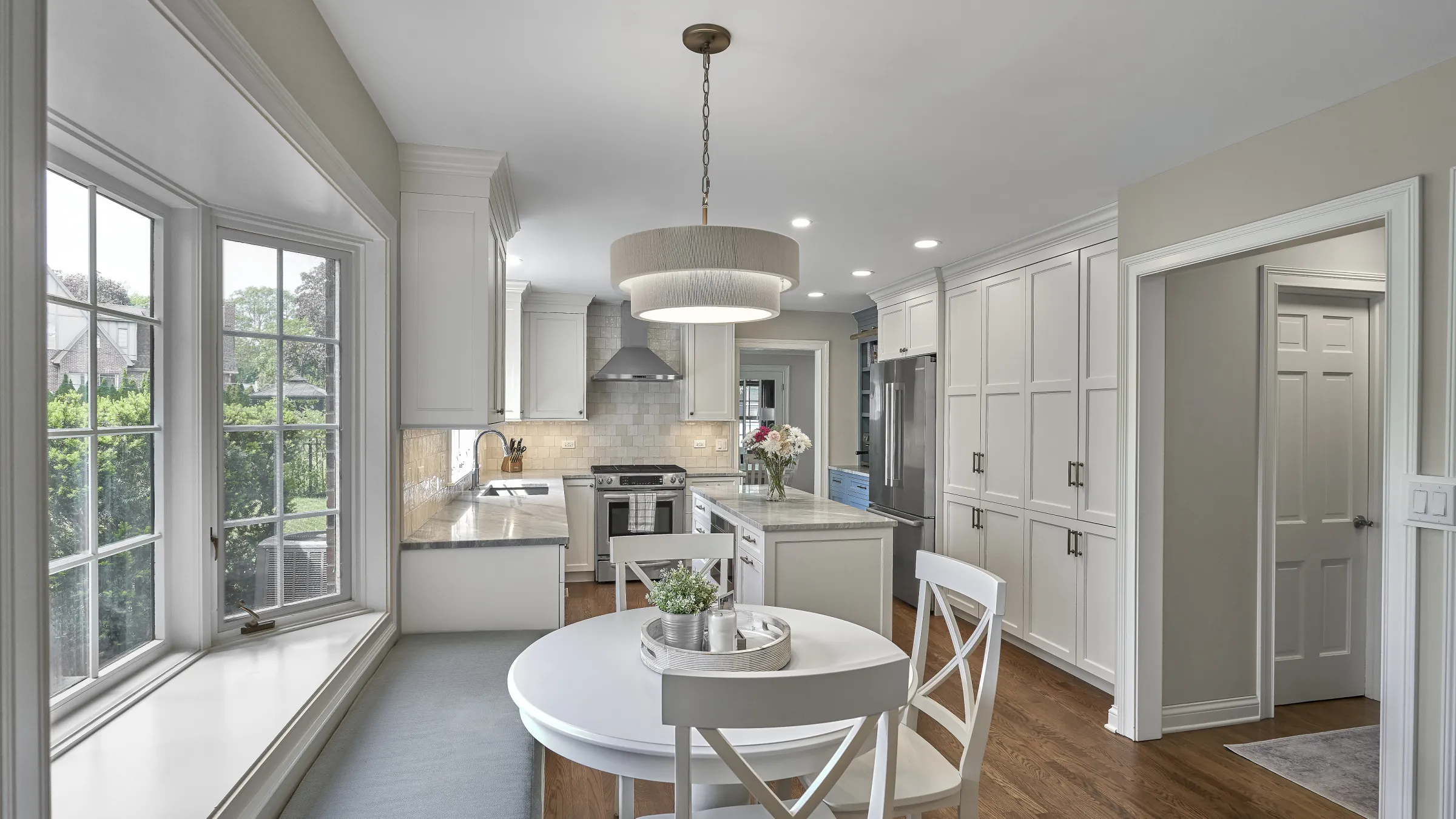 Modern white kitchen with shaker cabinets, marble countertops, stainless steel appliances, wood floor, and bay window seating