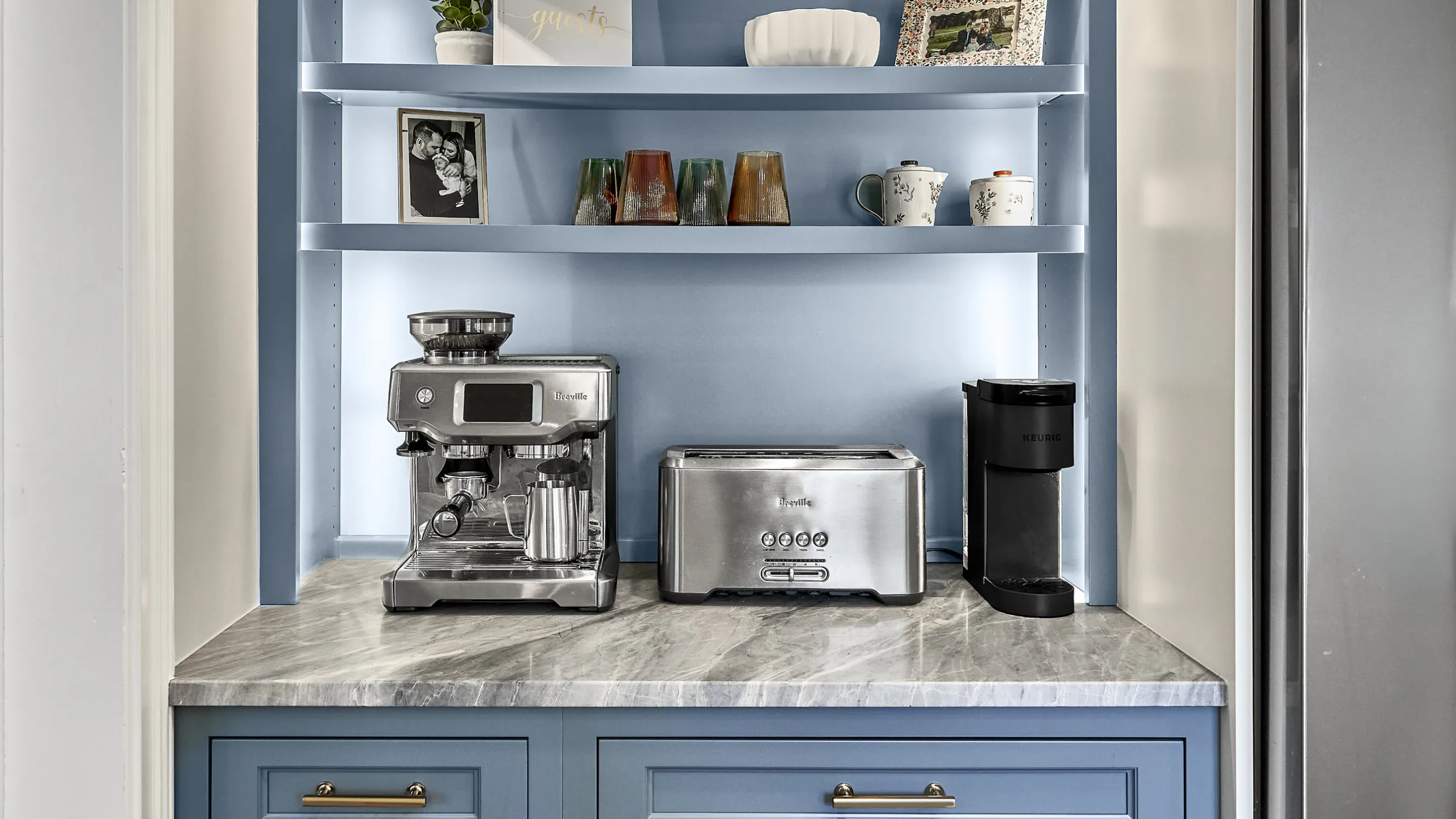 Blue kitchen cabinets with marble countertops, built-in shelving, espresso machine, toaster, and coffee maker.